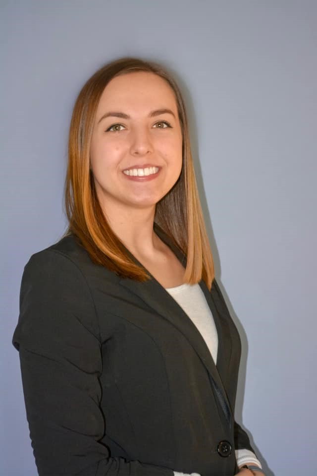 A young woman in a business suit, with a confident smile against a grey background.